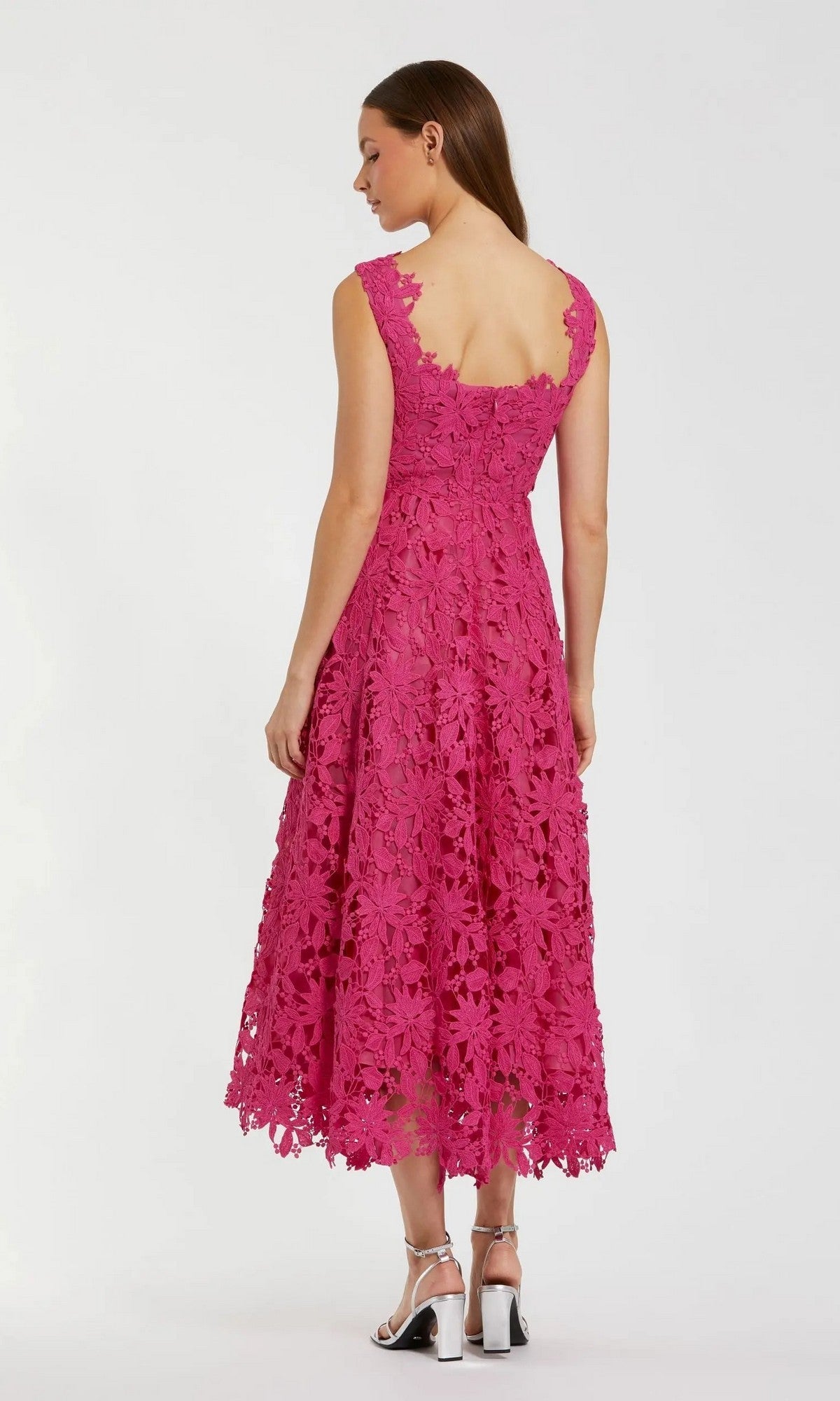 A woman stands facing away in front of a plain white background, wearing the Mac Duggal 69073 Midi Party Dress, a sleeveless, knee-length pink floral lace party dress, styled with silver sandals.